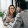 Two corporate business people sitting at a desk talking with a laptop on the table.